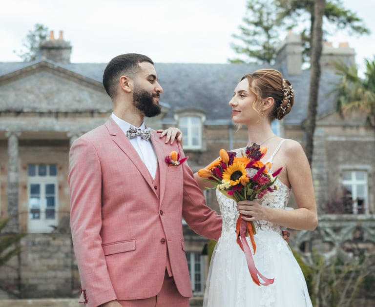 Une mariée et un marié se tiennent devant un manoir élégant, vêtu de leurs habits de mariage