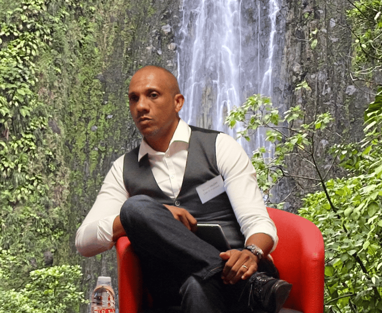 Bruno Valery lors d’une conférence sur le tourisme en Guadeloupe, avec les chutes du Carbet en arrière-plan.