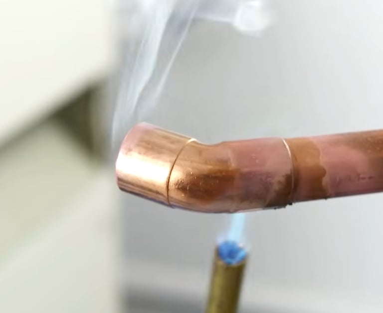 Professional plumber soldering a copper pipe elbow joint with a blue propane torch flame and rising smoke.