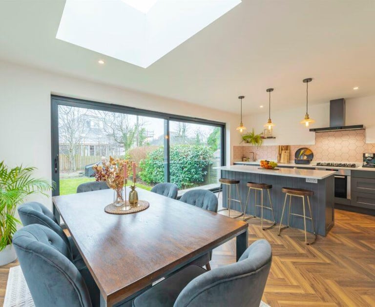 Modern open plan kitchen and dining room with herringbone floors, large skylight, and sliding glass doors.