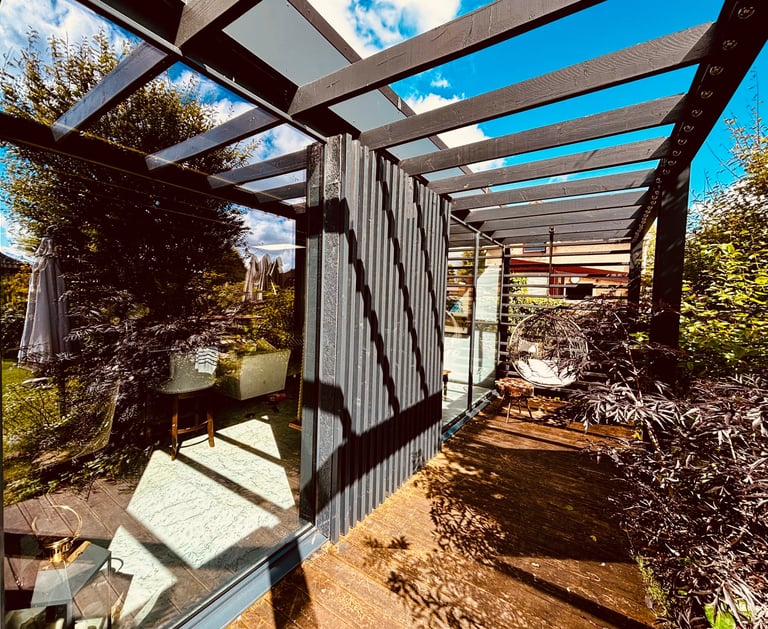 Modern garden pergola with a wooden deck, glass walls, and a hanging chair in a lush backyard.