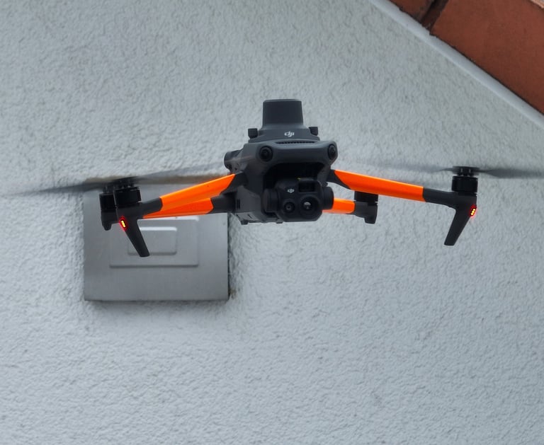 A DJI Mavic 3 Enterprise drone with orange arms hovering near a building wall for a roof inspection.