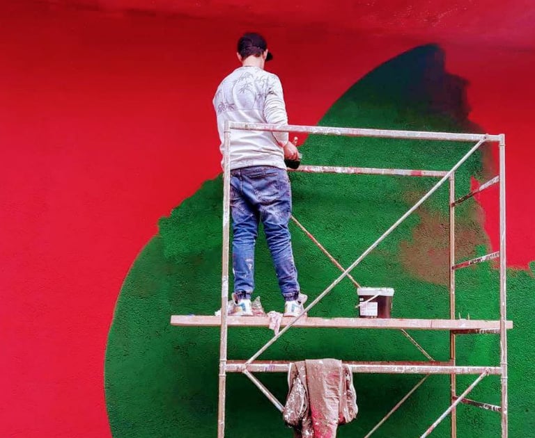 Mural painting in progress on a red and green wall, artist Poliops working from scaffolding