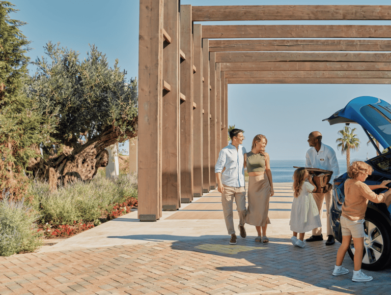 A family arrives at a luxury ikos resort as a hotel porter helps unload luggage from their blue car.
