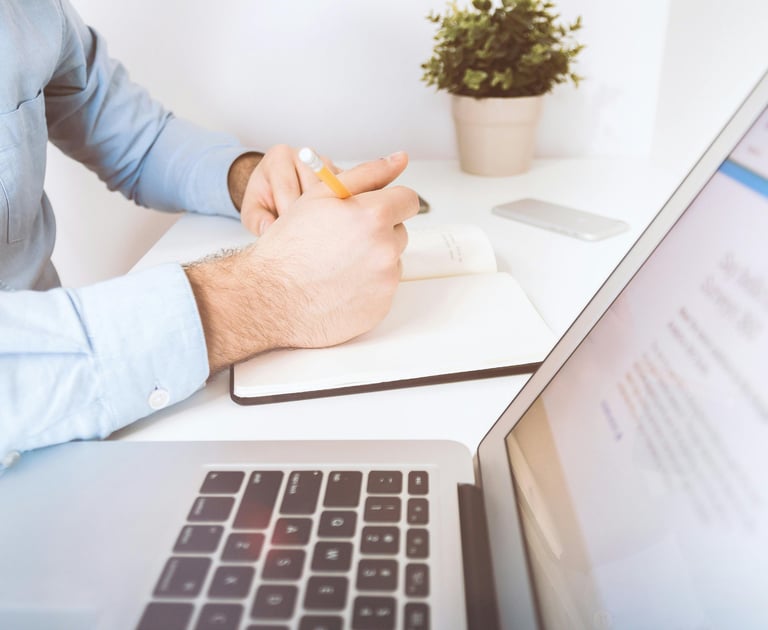 A desk with a laptop, open notebook, and a male writing in the notebook.