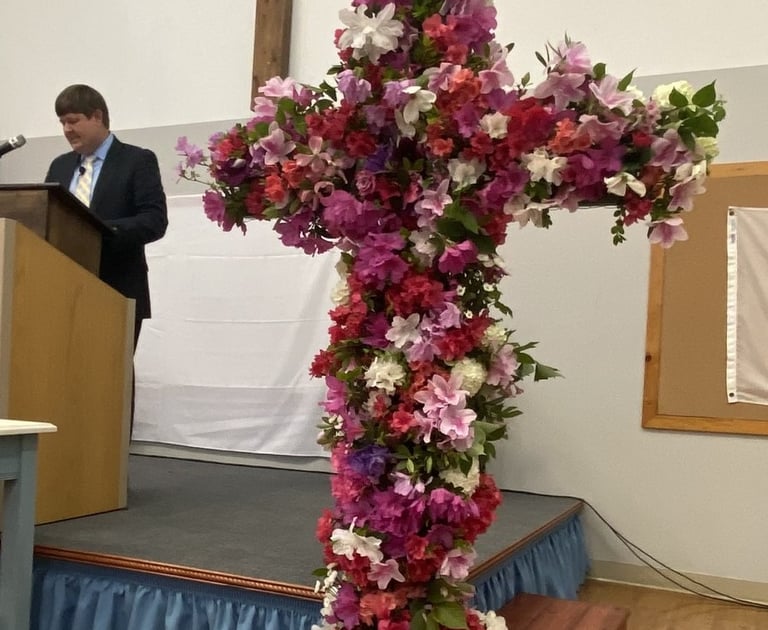 Pastor Wesley Daniels in the pulpit speaking and a large cross covered in Easter flowers