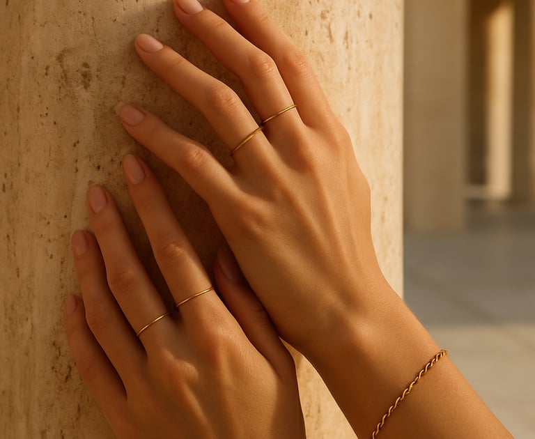 Close-up editorial photograph of a woman's hands wearing delicate gold minimalist rings and a classic chain bracelet, gently touching a warm sand-toned travertine stone column. Soft, warm natural light, high-end fashion catalog style, set in a modern Western European open-air space.