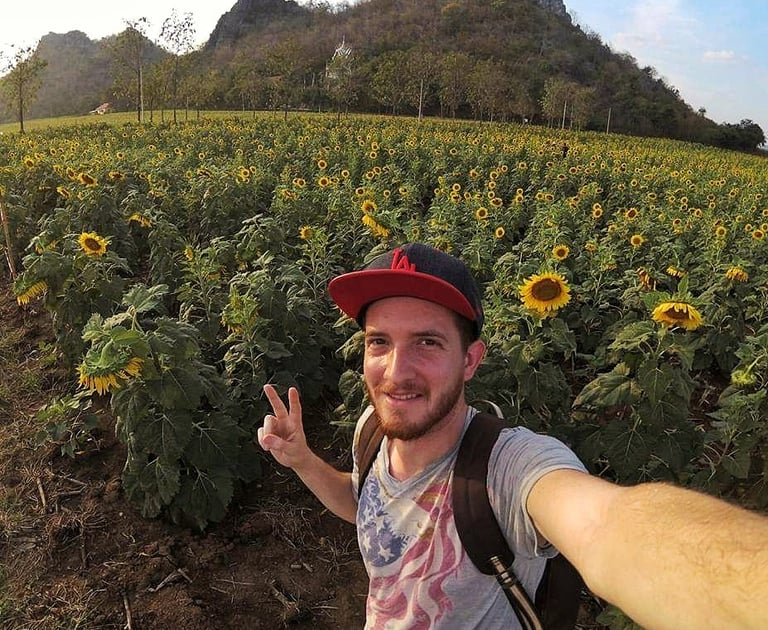 Smiling taking a selfie in a blooming sunflower field with mountains in the background.