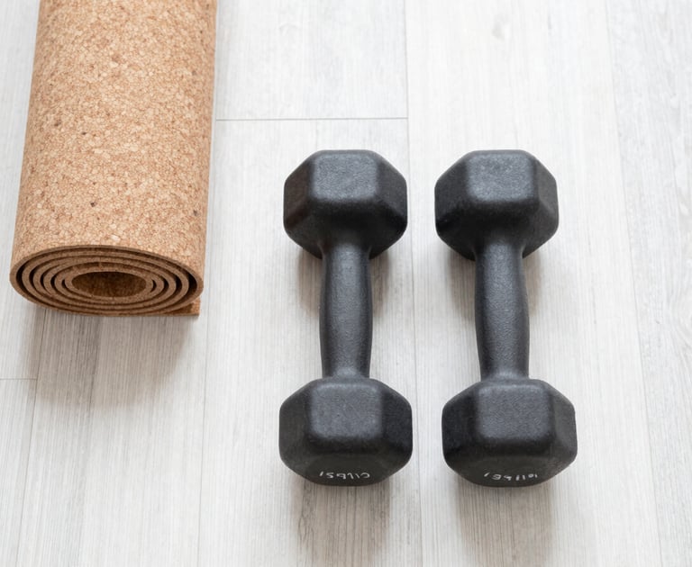 Top-down flat lay of a matte black dumbbell and a cork yoga mat on a bright white oak floor, soft morning daylight, airy neutral tones, 35mm