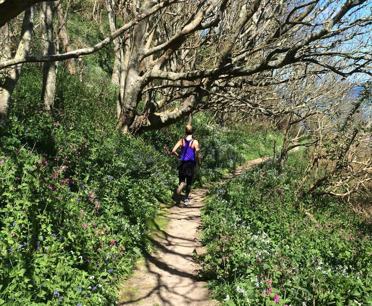 A woman trail running on a sunny path through a lush green forest with blooming wildflowers.