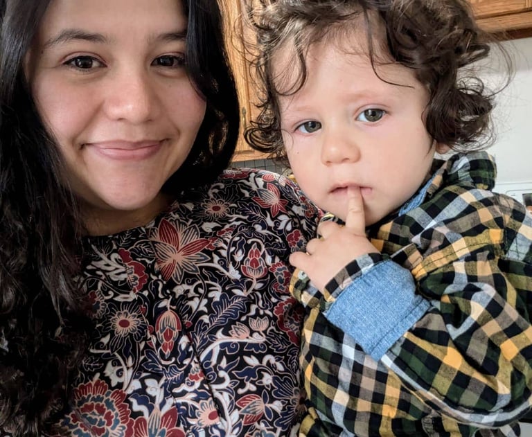 Smiling woman holding a curly-haired toddler wearing a plaid shirt in a home kitchen setting.