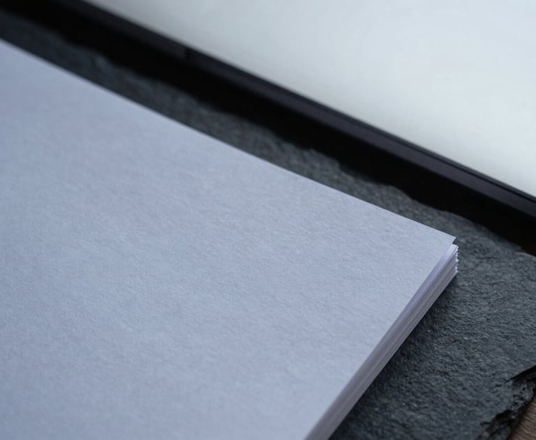 Close-up of heavy textured paper documents resting on a dark slate desk, architectural shadows, cool overcast daylight.
