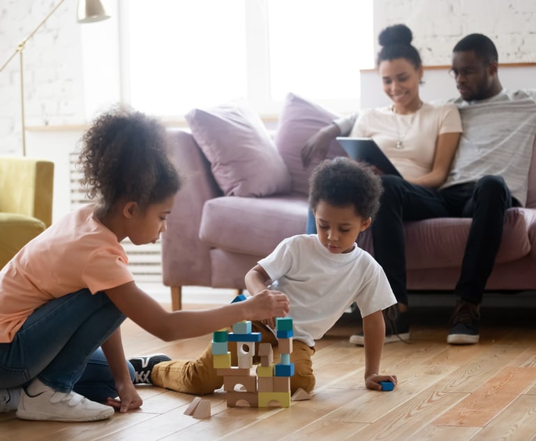a girl and boy playing with blocks on the floor with parents planning in the background