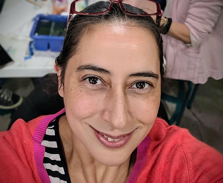 A smiling woman with brown eyes and hair wearing a bright pink cardigan in an art studio.