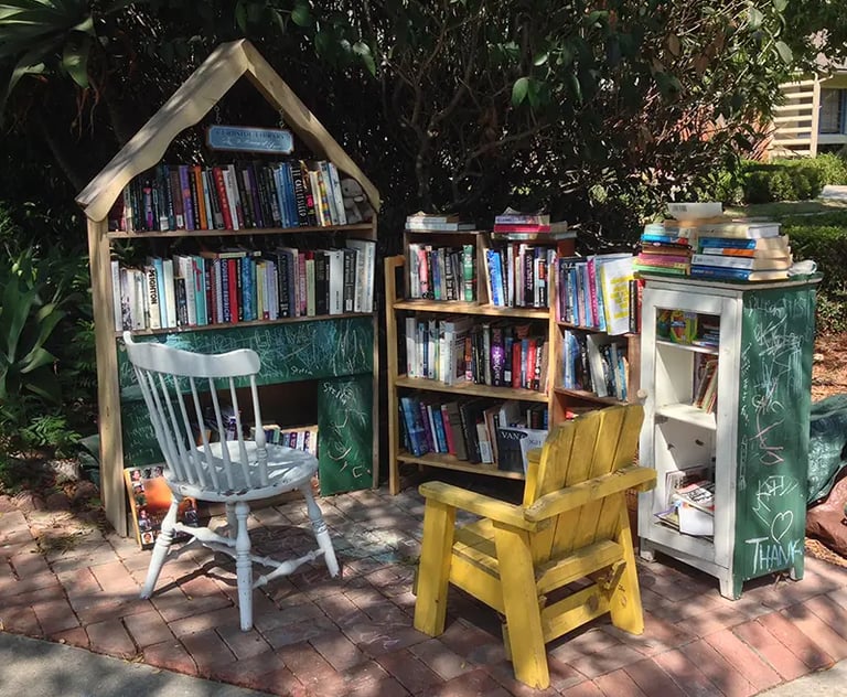 Outdoor community library on a brick patio featuring wooden bookshelves, a white wooden chair, and a yellow chair.
