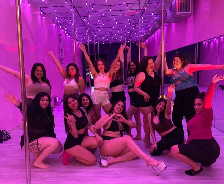 Twelve people posing together in a pole dance studio lit with a pink light