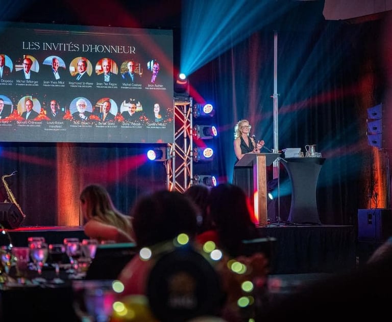 A woman speaking at a gala podium next to a digital screen showing guests of honor.