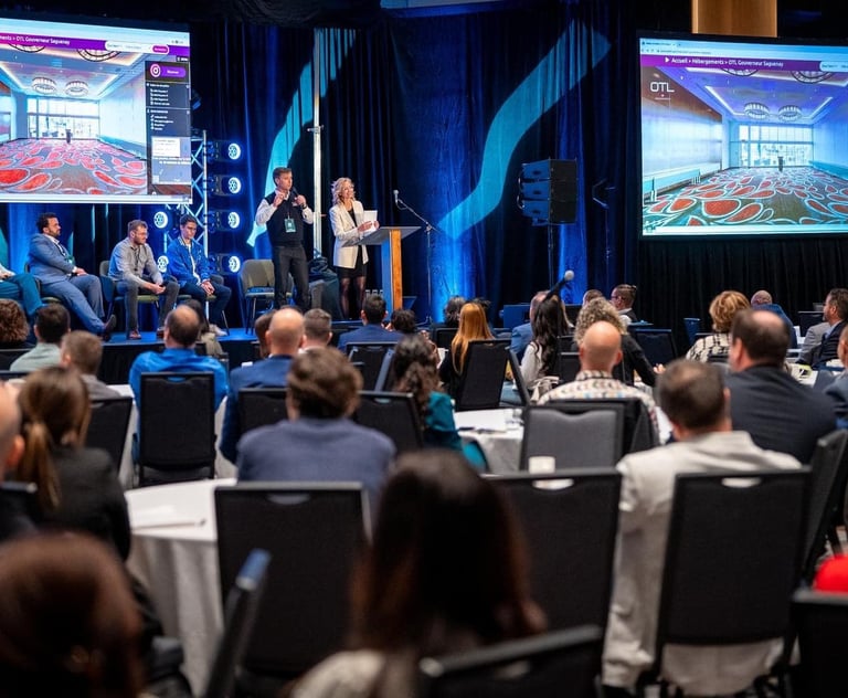 Speakers present at a corporate conference panel on stage with large digital screens for the audience.