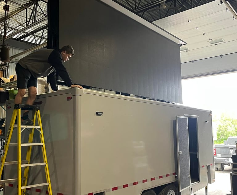 A technician installs a large mobile LED screen on a white custom trailer inside a warehouse.