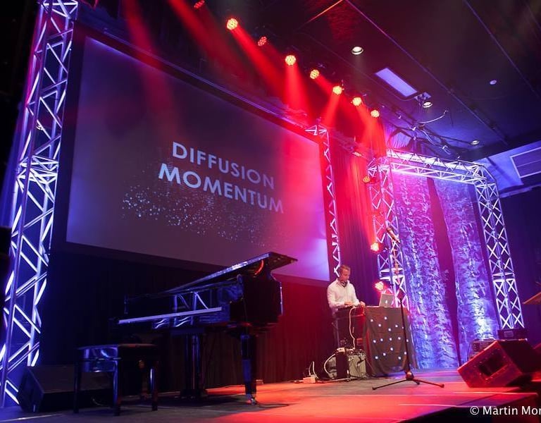 A grand piano on a stage with red and blue concert lighting and a Diffusion Momentum screen.
