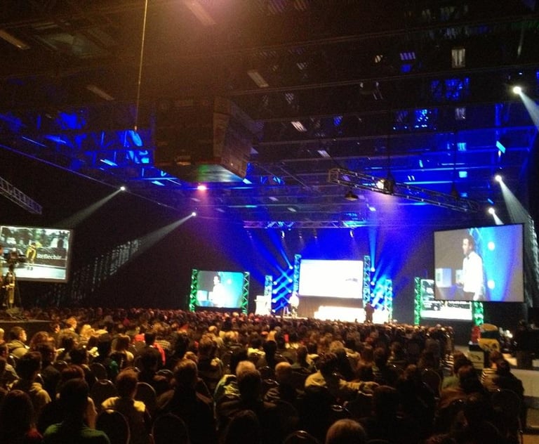 Crowd at a large tech conference with blue stage lighting and large video screens.