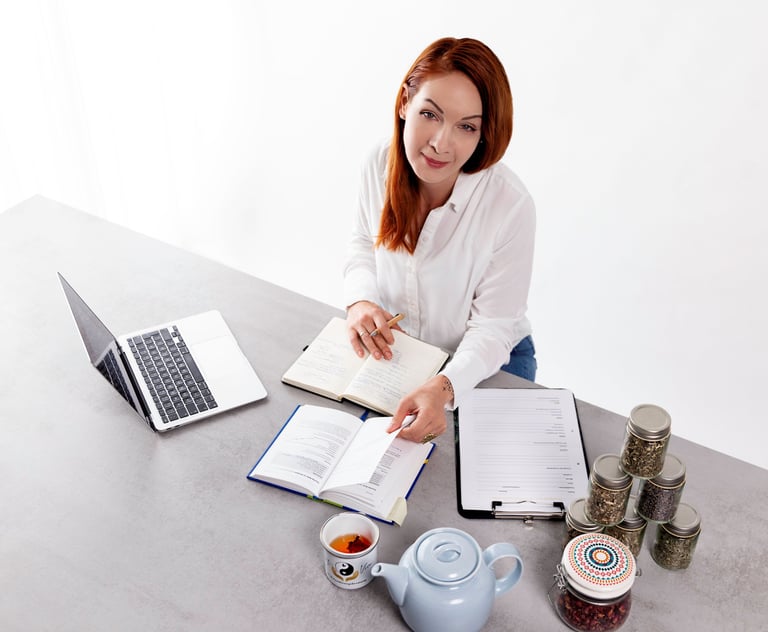 A nutritionist with red hair studying herbal tea recipes and jars of dried herbs at a desk.