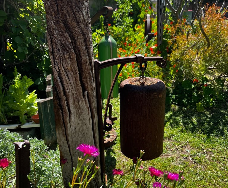 Rustic garden bells hanging from a wooden post amidst vibrant pink flowers and lush greenery.