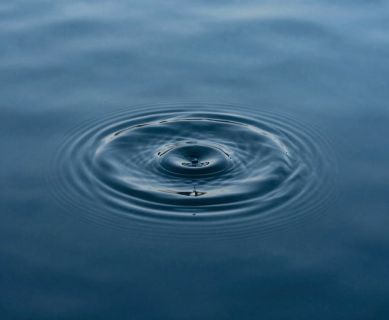 A minimalist photograph of a single ripple expanding on the surface of a deep steel blue pool of water. The composition is tight and focused, emphasizing the profound stillness of the surrounding liquid. Soft, diffuse lighting.