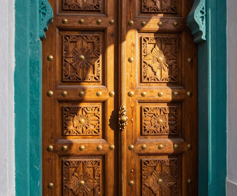 Close architectural view of a tall carved wooden Moroccan riad door, deep teal-painted frame with intricate geometric brass nailheads, warm golden-hour sidelight raking across the carved relief, shallow depth of field, left edge kissing the frame