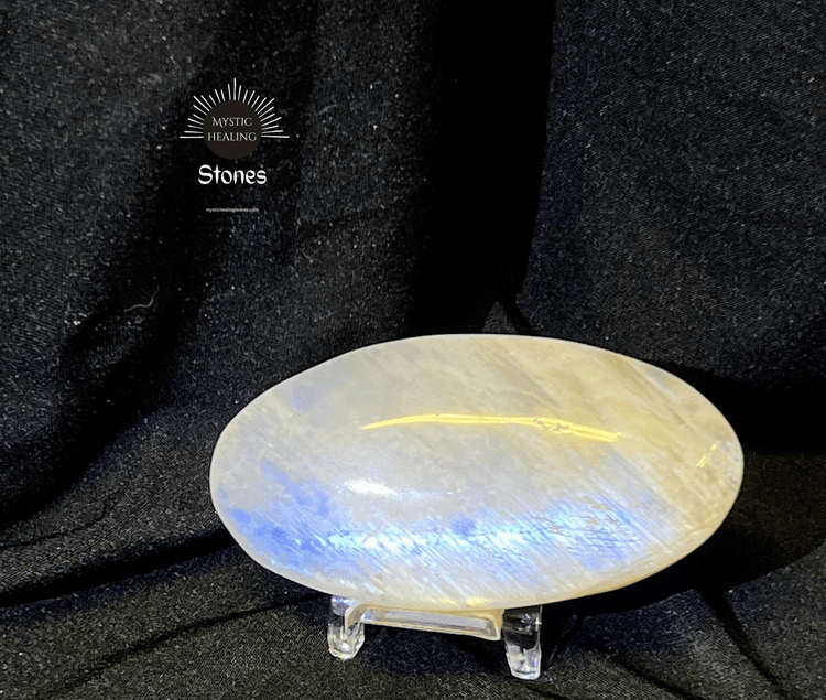 Polished oval moonstone palm stone with blue flash on a stand against black fabric.
