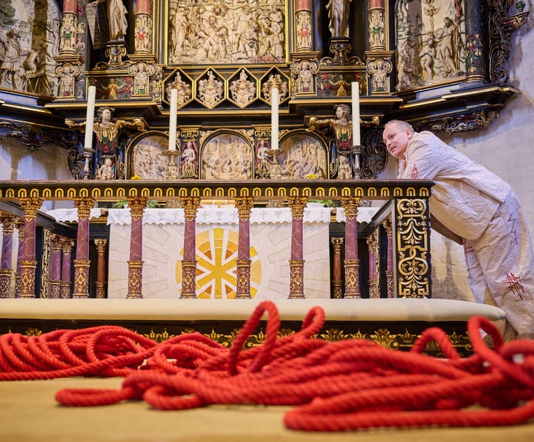 Ein Schauspieler stützt sich auf einen Kirchenaltar. Im Vordergrund ein langes rotes Seil.