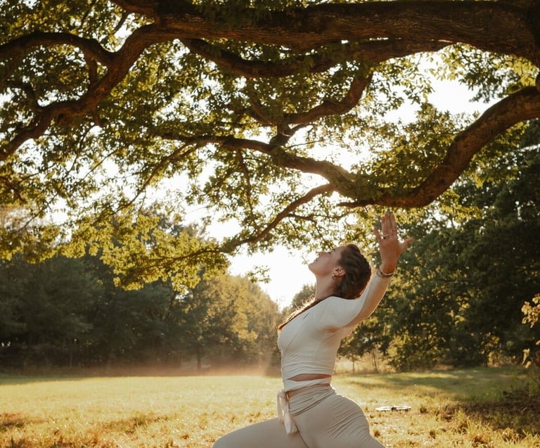 Eine Frau praktiziert Yoga im Krieger-1-Stand unter einem großen Baum in einer sonnigen Waldlichtung