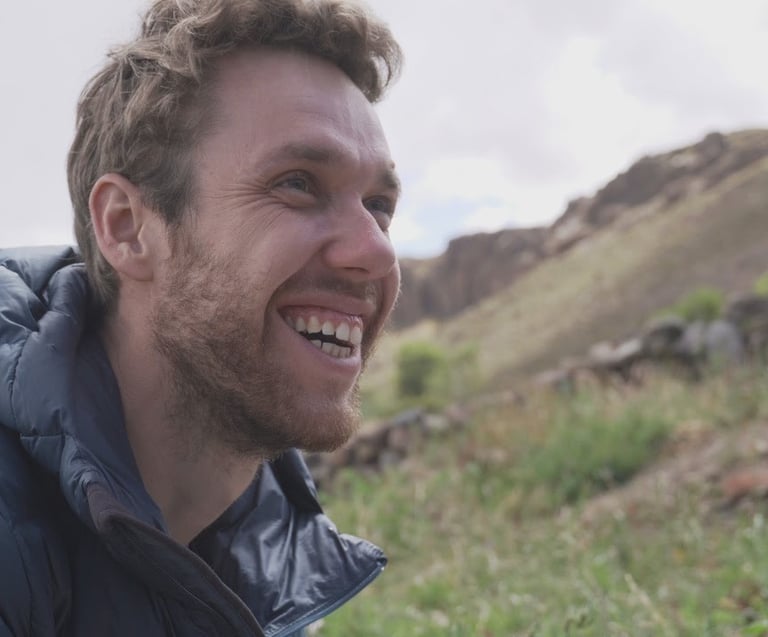 Un homme souriant, vêtu d'une doudoune bleue, en randonnée dans un paysage montagneux.