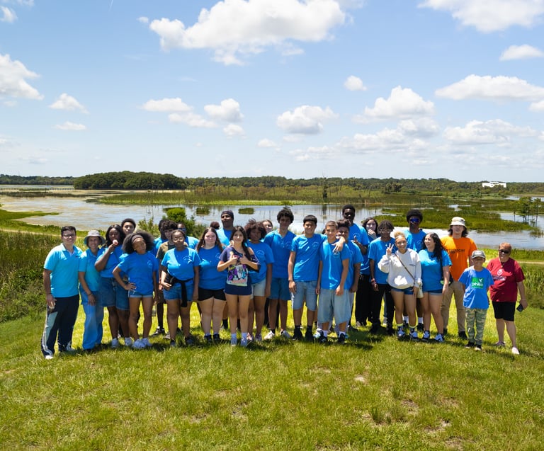 A diverse group of students in blue shirts posing for a photo at a scenic Florida nature preserve wetland.