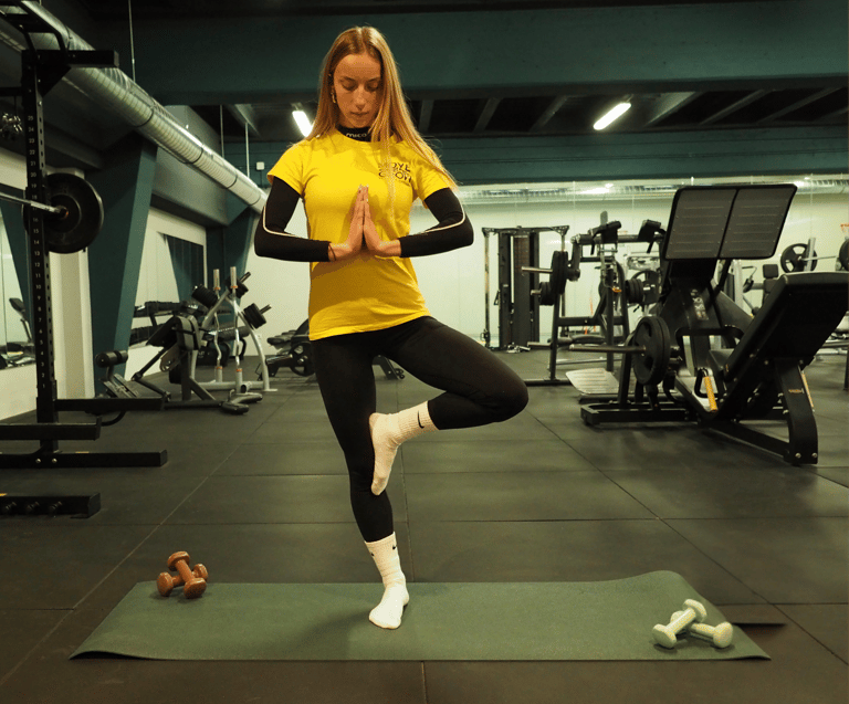 Una donna in posa di yoga albero (Vrksasana) su un tappetino da yoga in palestra. Indossa una maglietta gialla con logo 'Move