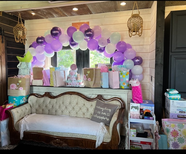 Purple and white balloon arch over baby shower gift table and vintage tufted sofa.