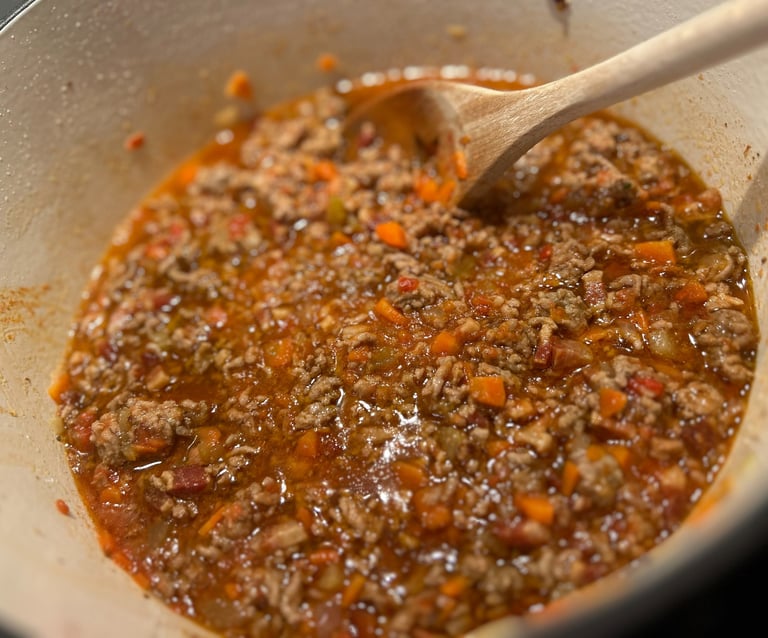 A wooden spoon stirs a savory bolognese meat sauce simmering with carrots in a white Dutch oven.