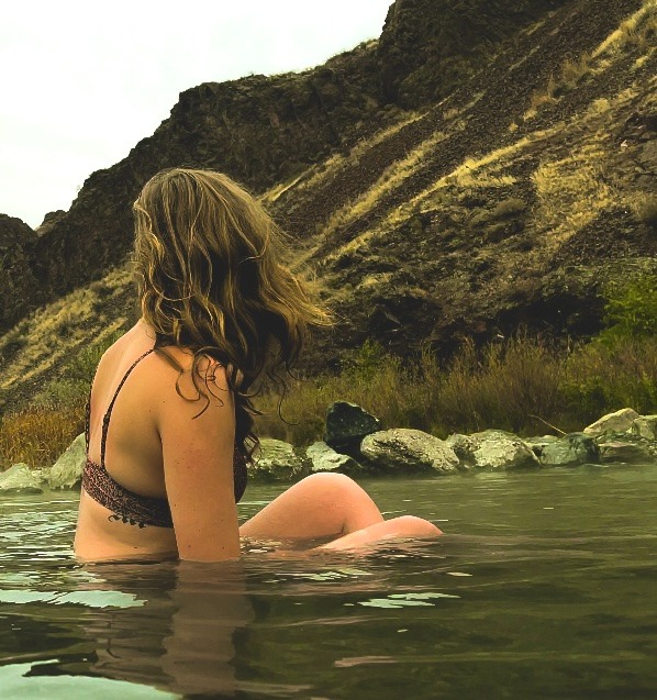 A woman with wavy hair relaxing in a natural hot spring pool with rocky cliffs in the background.