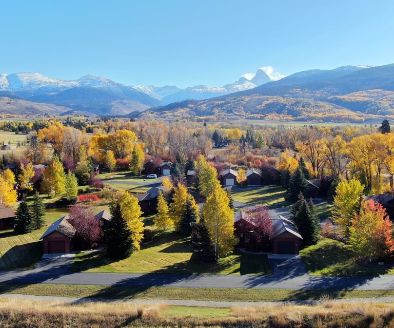 Aerial view of a mountain valley neighborhood with autumn foliage and snow-capped peaks in the background.