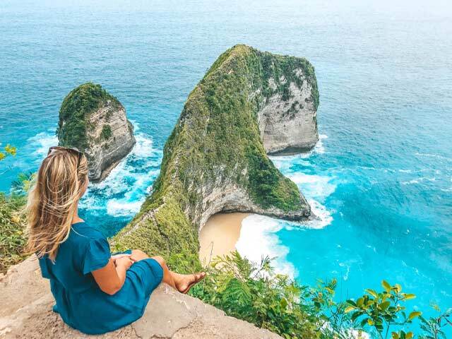 A woman sits overlooking Kelingking Beach in Nusa Penida, Bali, with turquoise water and cliffs.