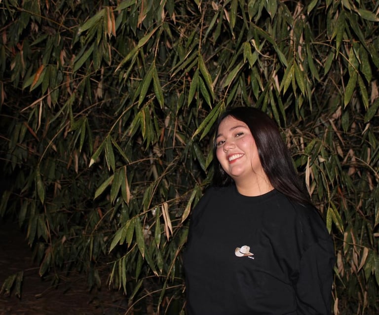 A smiling young woman in a black t-shirt posing in front of lush green bamboo leaves at night.