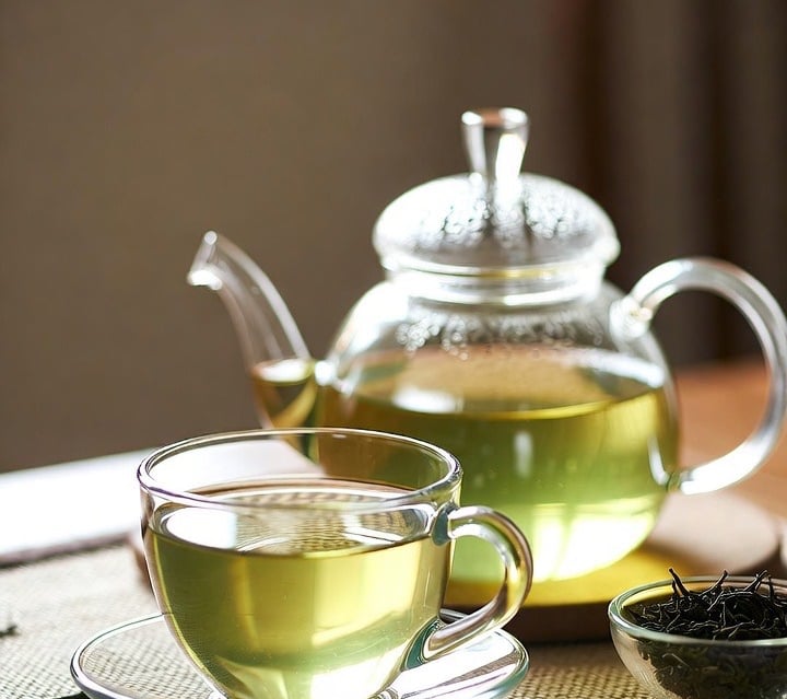 Freshly brewed green tea in a clear glass cup and teapot on a bamboo mat.