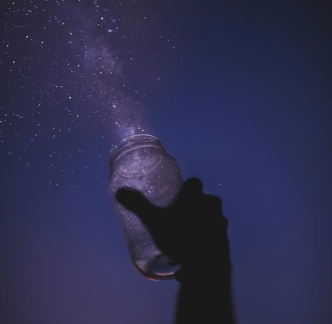 Silhouette of a hand holding a glass jar catching a purple Milky Way galaxy and stars.