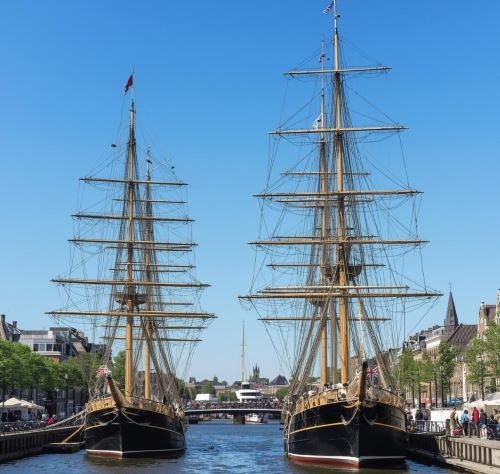 Two historic tall ships with wooden masts docked in a city canal under a clear blue sky.