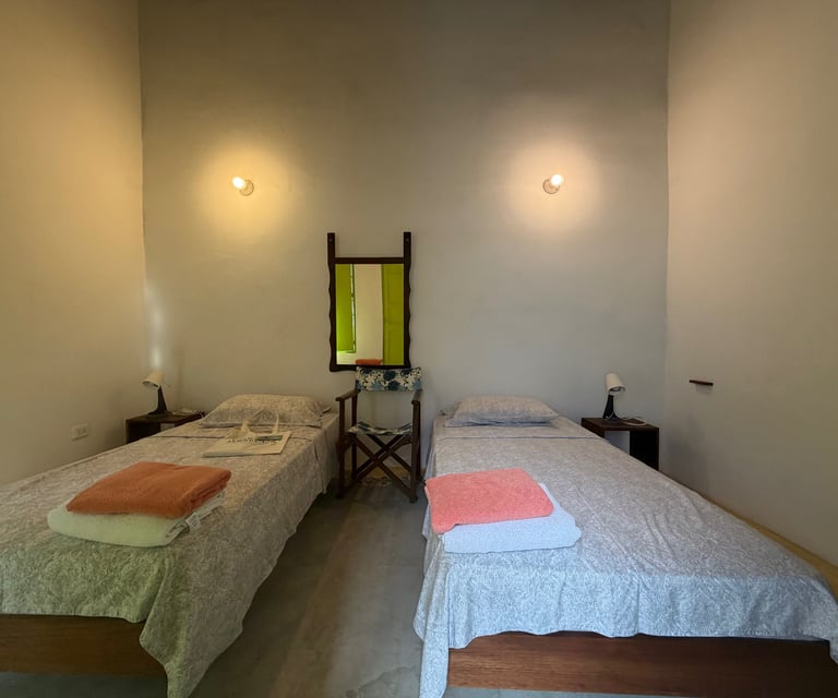 A minimalist guest room featuring two twin beds, bedside lamps, and a folding chair below a wall mirror.