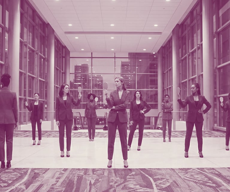 A diverse group of women in colored business suits strike a pose, holding up hand mirrors