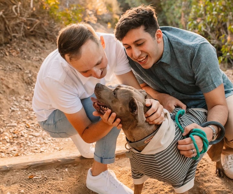 Two people crouch on a dirt path, affectionately petting a dog in a striped sweater.