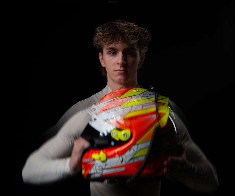 Young race car driver holding a vibrant orange and yellow racing helmet against a dark background.