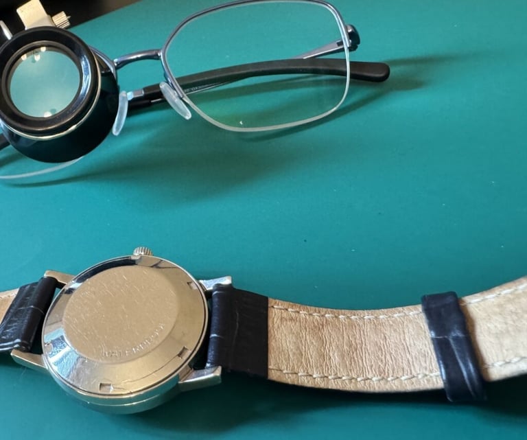 Vintage watch back, jeweler's loupe, and eyeglasses on a green surface for watch repair.
