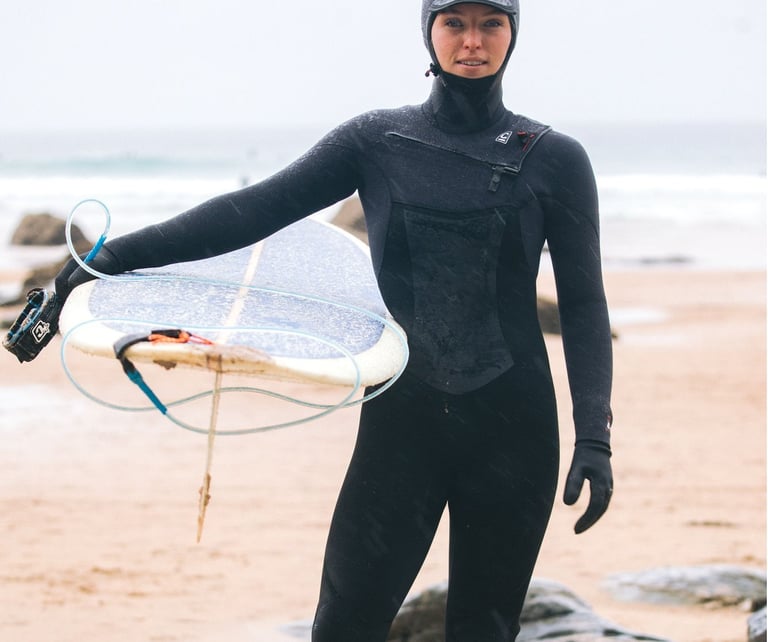 A female surfer wearing full winter kit holding a longbaord at the beach